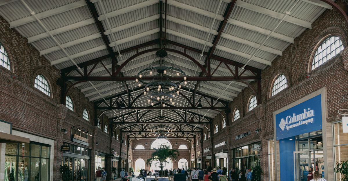 Urban shopping mall interior with people walking amid stores under decorative lighting and exposed beams.